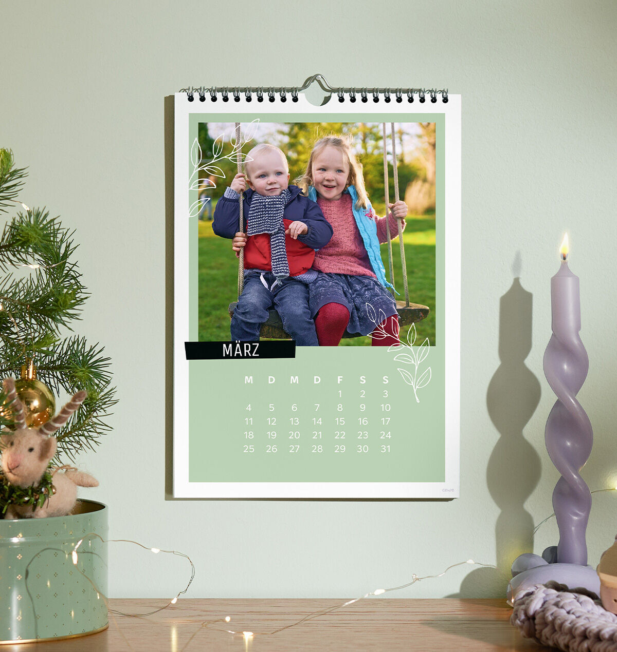 Personalisierter CEWE Fotokalender an der Wand mit einem Bild von zwei Kindern auf einer Schaukel, daneben eine Kerze und Weihnachtsdekorati