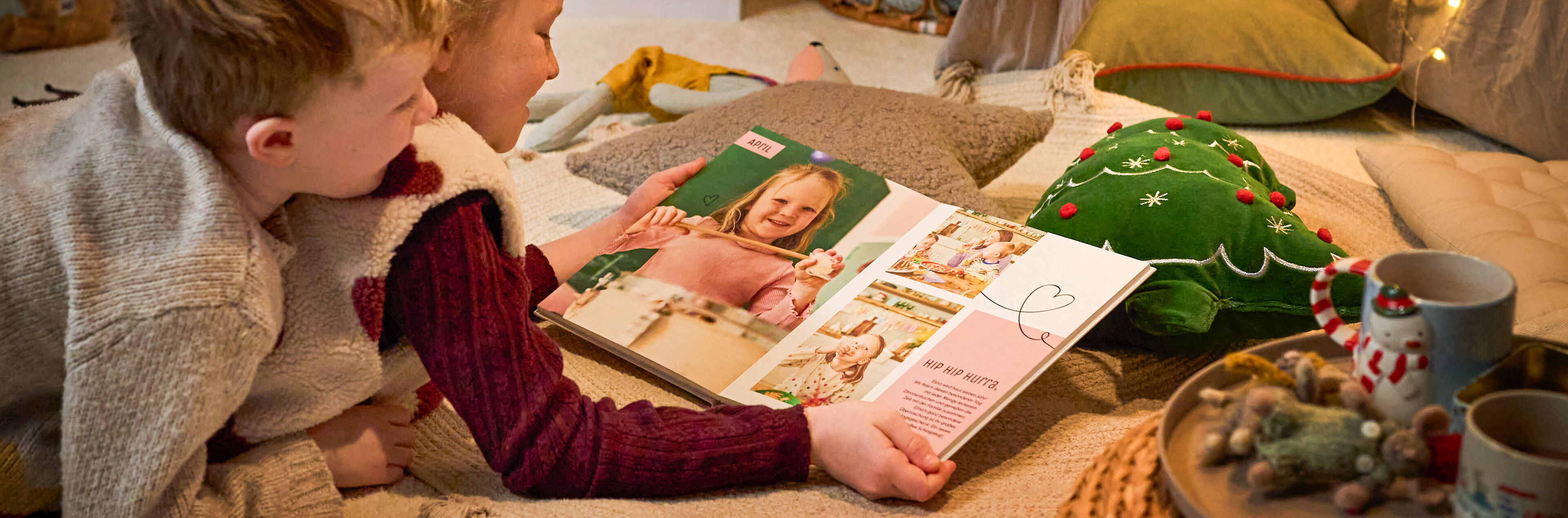 Zwei Kinder betrachten gemeinsam ein CEWE FOTOBUCH mit Familienfotos in weihnachtlicher Umgebung.