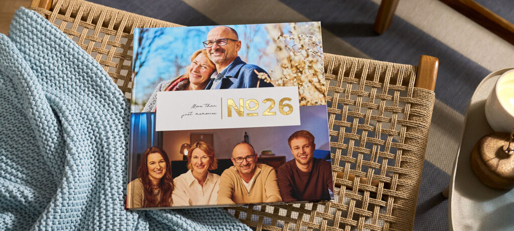 CEWE FOTOBUCH „N°26“ mit Fotos eines Paares und einer Familie von vier Personen auf einem geflochtenen Stuhl mit blauer Decke.