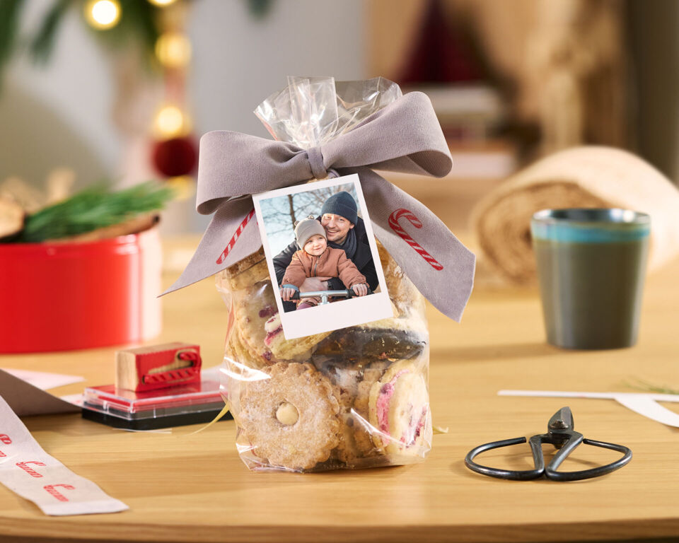 Geschenktüte mit Keksen, Schleife und Fotokarte auf einem Tisch, umgeben von Bastelzubehör.
