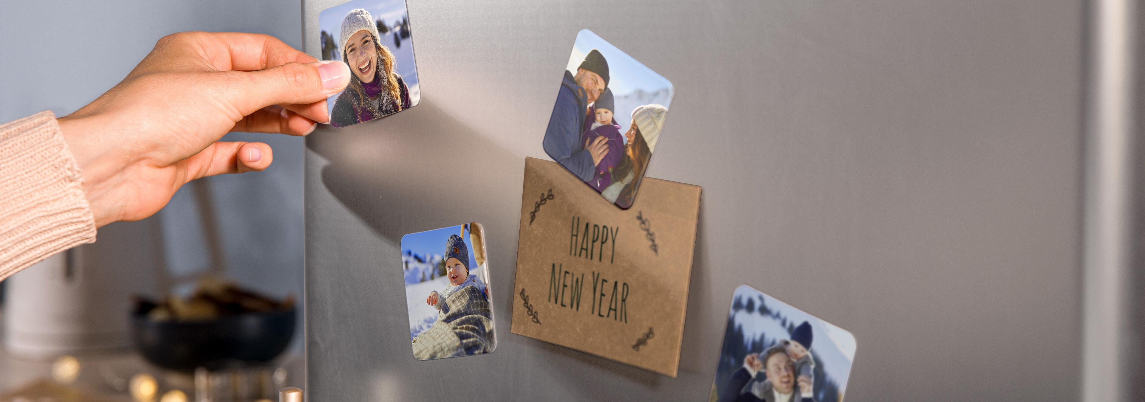 Person befestigt personalisierte Fotomagnete mit winterlichen Familienfotos an einem Kühlschrank neben einer Karte mit der Aufschrift „Happy