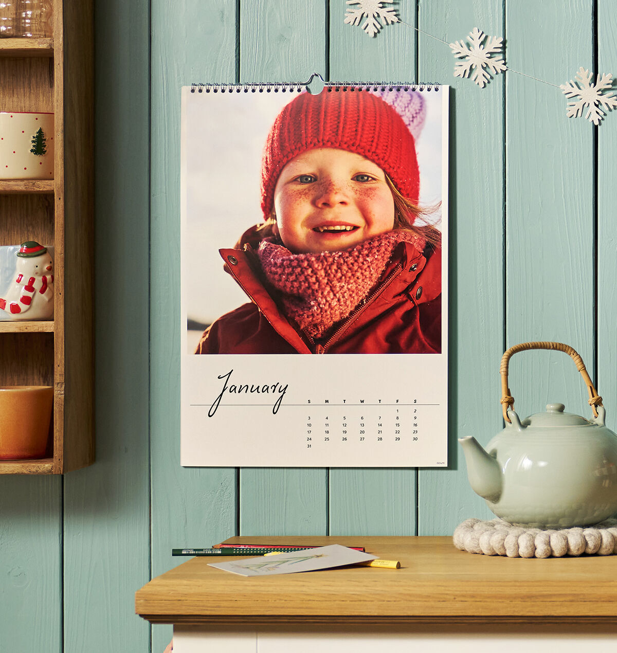 CEWE Wandkalender mit winterlichem Kinderfoto an türkiser Holzwand, daneben ein Tassenregal und eine Teekanne.
