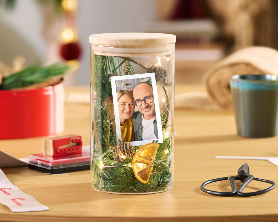Dekoratives Glas mit Holzdeckel, Tannengrün, Lichterkette, Orangenscheibe und dem Foto eines Paares.