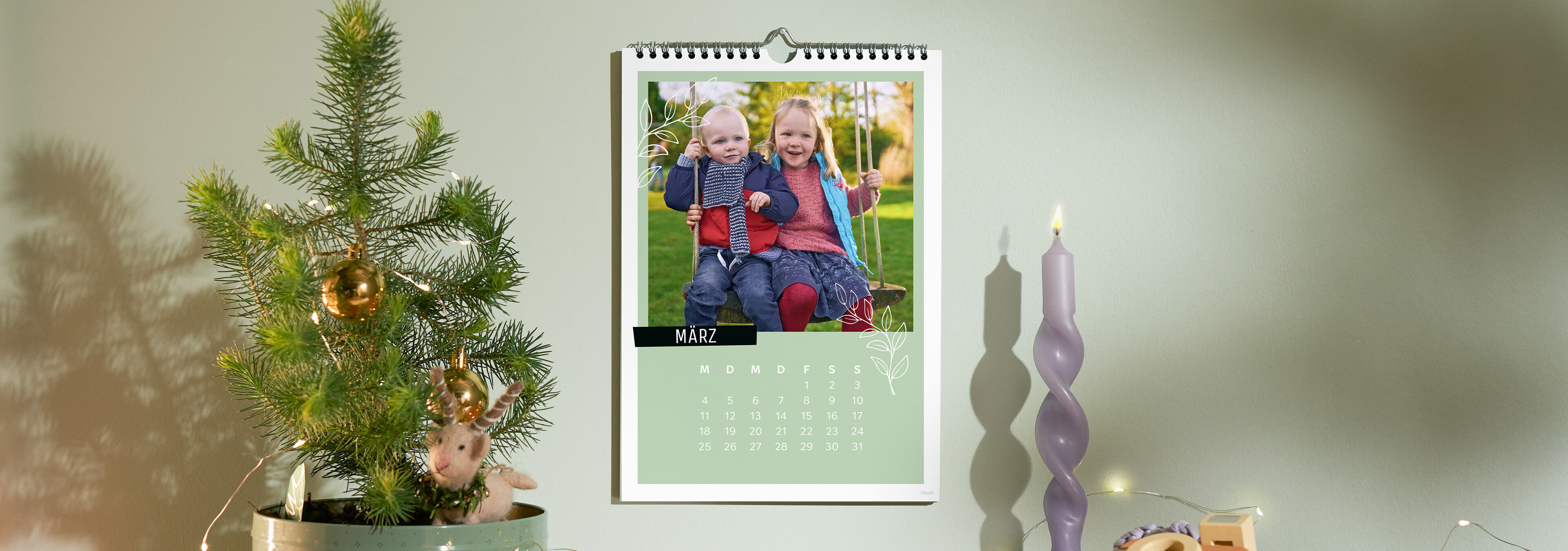 Personalisierter CEWE Fotokalender an der Wand mit einem Bild von zwei Kindern auf einer Schaukel, daneben eine Kerze und Weihnachtsdekorati