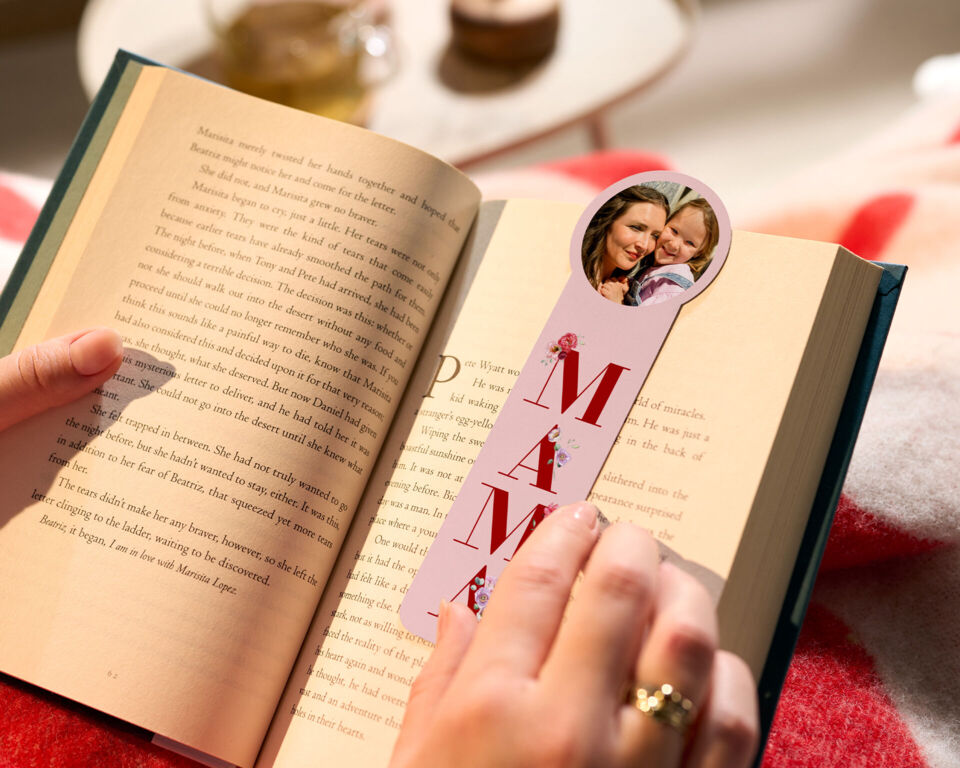 Hand hält ein Buch und ein Foto-Lesezeichen mit einem Foto von Mutter und Kind und dem Schriftzug "MAMA".