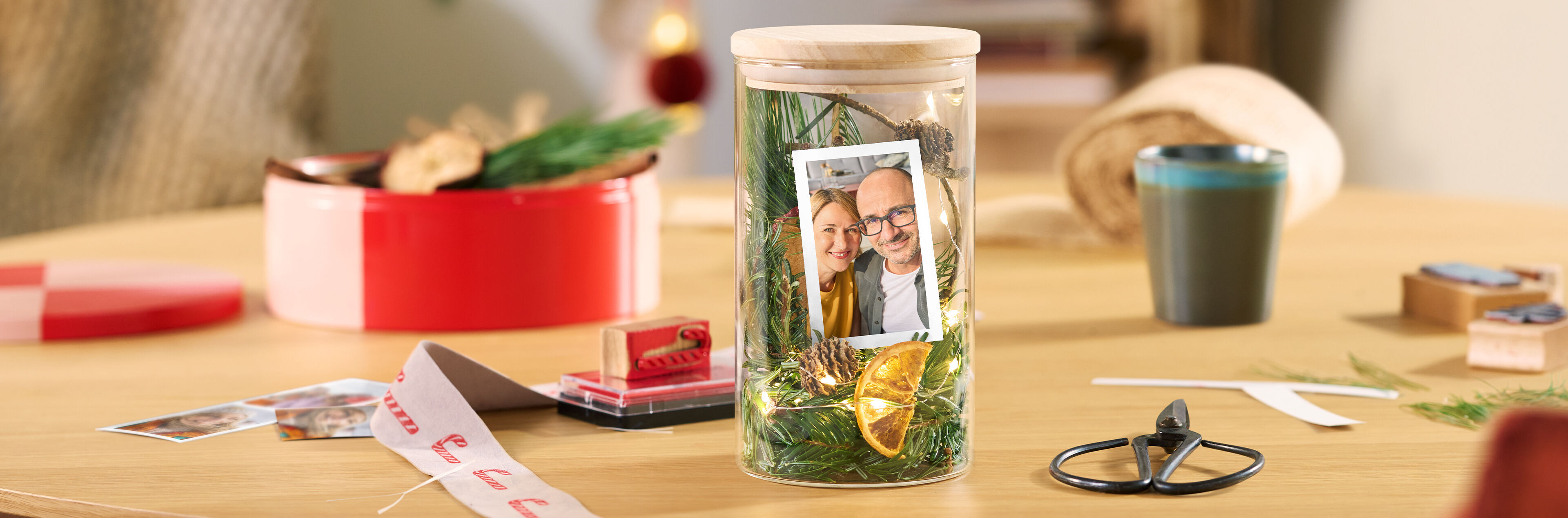 Dekoratives Glas mit Holzdeckel, Tannengrün, Lichterkette, Orangenscheibe und dem Foto eines Paares.