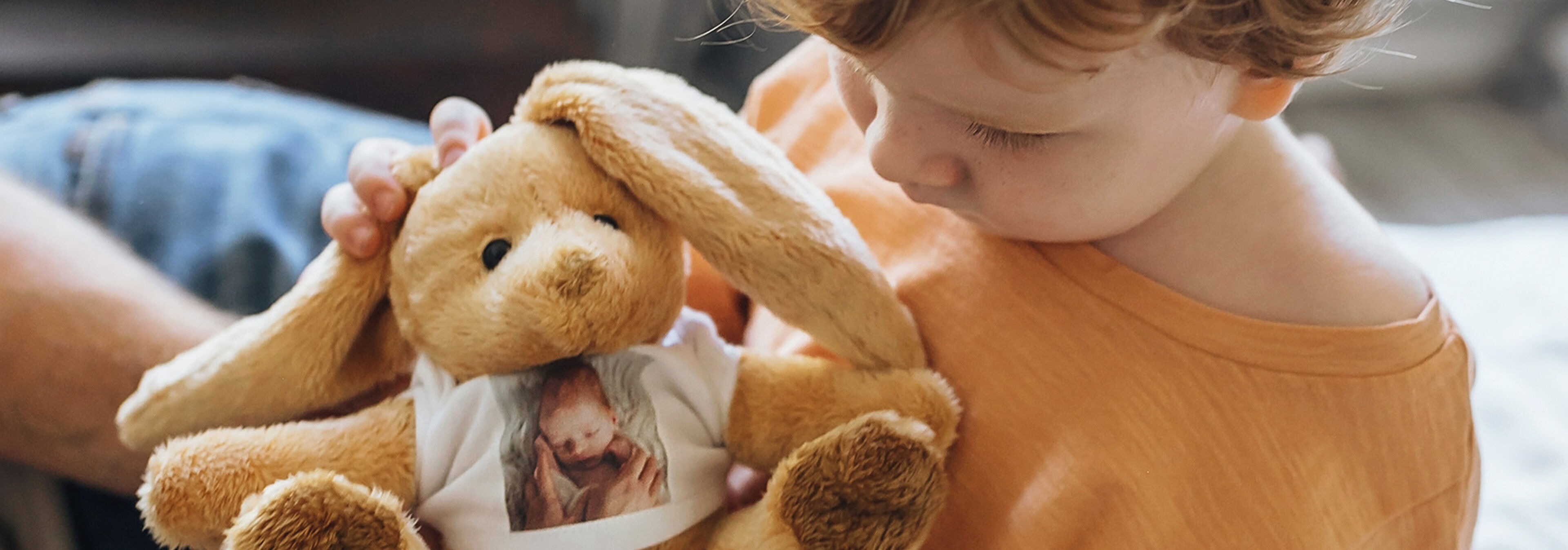 Kind hält einen Kuschelhasen mit Babyfoto auf dem T-Shirt in den Armen.