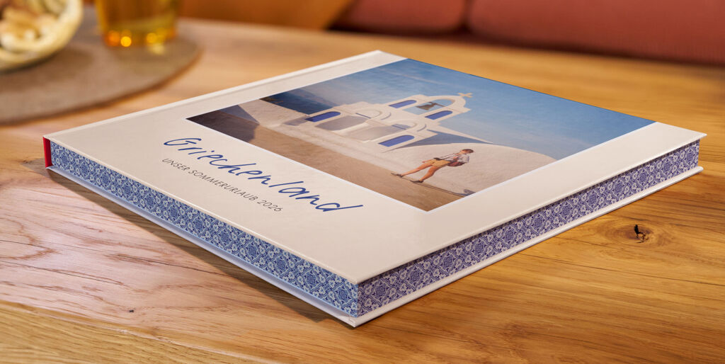 Auf einem Couchtisch liegt ein CEWE FOTOBUCH mit Farbschnitt, das Cover zeigt ein Urlaubsfoto und den Schriftzug „Griechenland“.