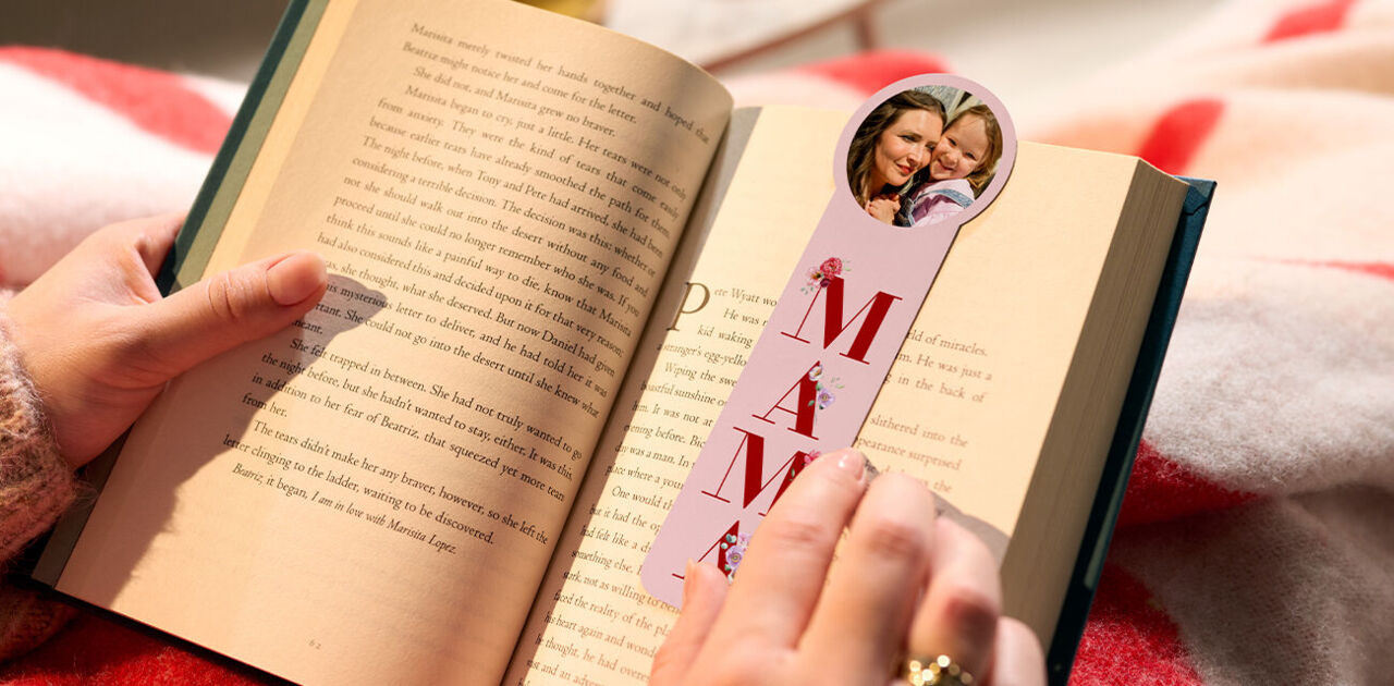 Hand hält ein Buch und ein Foto-Lesezeichen mit einem Foto von Mutter und Kind und dem Schriftzug "MAMA".