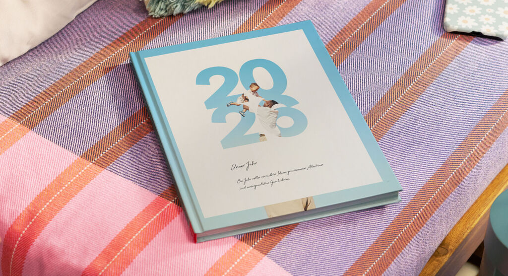 CEWE FOTOBUCH mit türkisfarbenem Einband und der Aufschrift „2026", das auf einer orange-pink gestreiften Decke liegt.