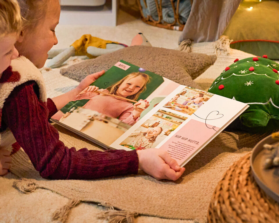 Zwei Kinder betrachten gemeinsam ein CEWE FOTOBUCH mit Familienfotos in weihnachtlicher Umgebung.