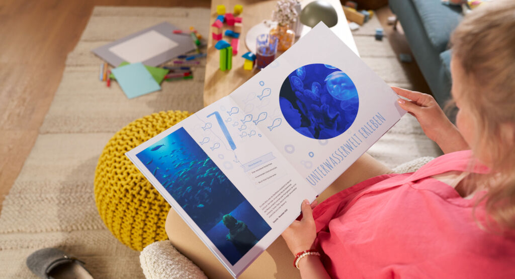 Eine Frau sitzt auf einem Sofa und blättert durch ein CEWE FOTOBUCH, die Doppelseite zeigt Fotomotive und die Zahl „7“.
