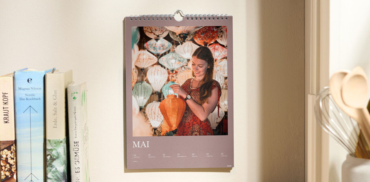 Ein CEWE Fotokalender hängt in einer gemütlichen Küche an der Wand, während eine Person lächelt.