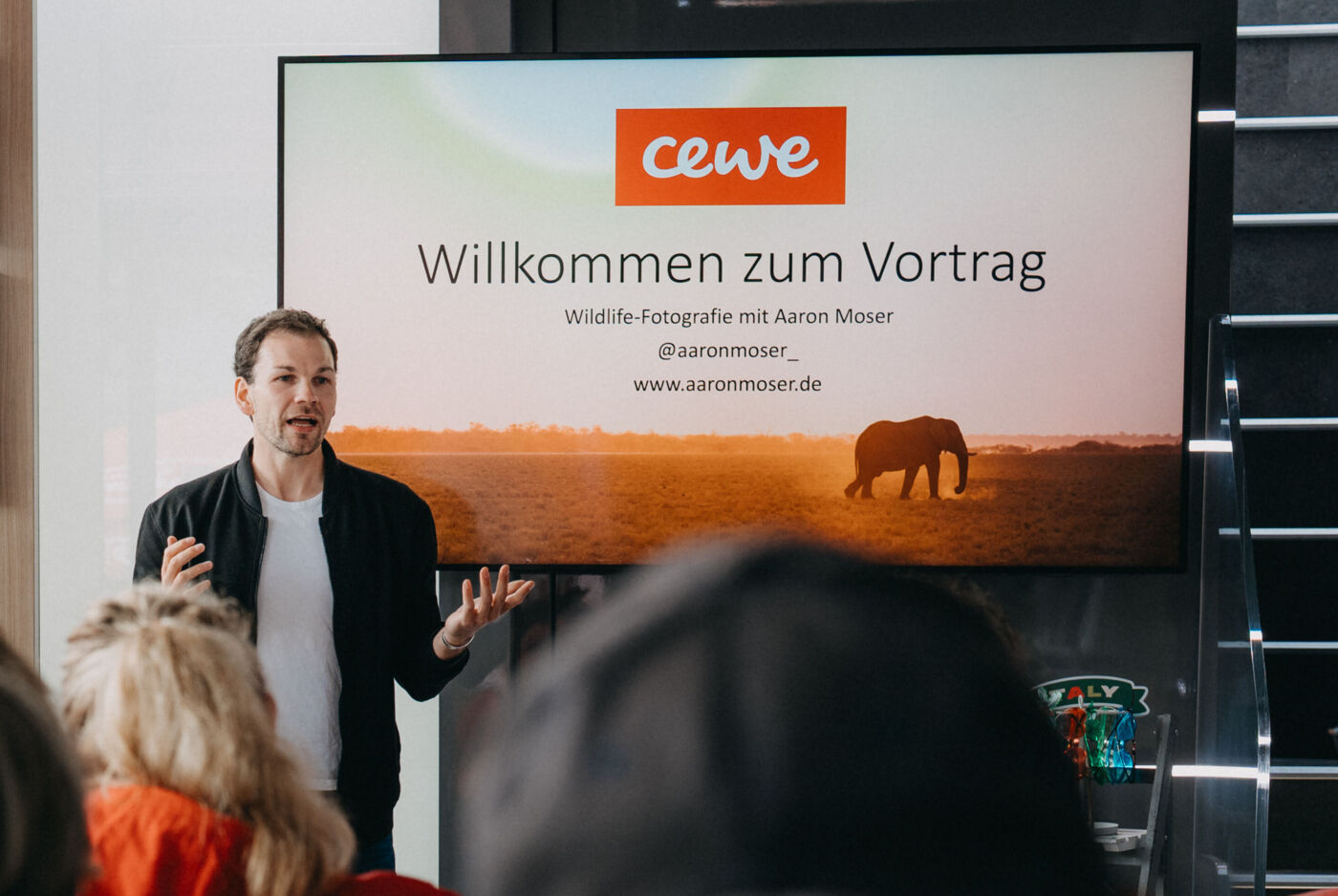 Ein Mann steht vor einem großen Bildschirm und hält einen Vortrag. Vor ihm sitzt ein Publikum in mehreren Reihen.