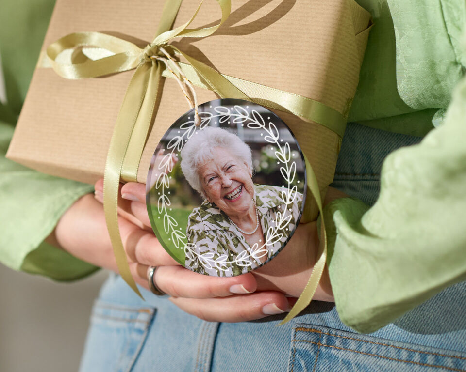 Person hält ein Geschenk mit Schleife und rundem Anhänger mit Foto einer lächelnden älteren Person hinter dem Rücken.