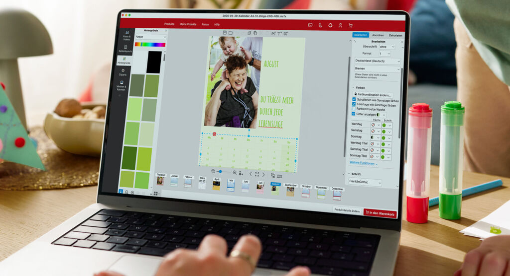 Eine Frau sitzt vor einem Laptop und gestaltet einen Fotokalender. Im Hintergrund ist ein spielendes Kind zu sehen.