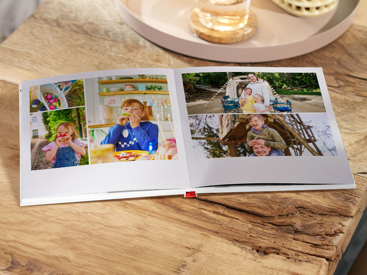 Aufgeschlagenes Fotobuch auf einem Holztisch mit mehreren Familienfotos: Kinder beim Essen und Spielen sowie gemeinsame Momente im Freien.