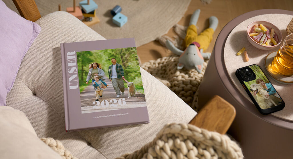 Auf einem Sofa liegt ein lila Fotobuch mit dem Titel „UNSER 2026“ und dem Foto einer Familie mit Hund.