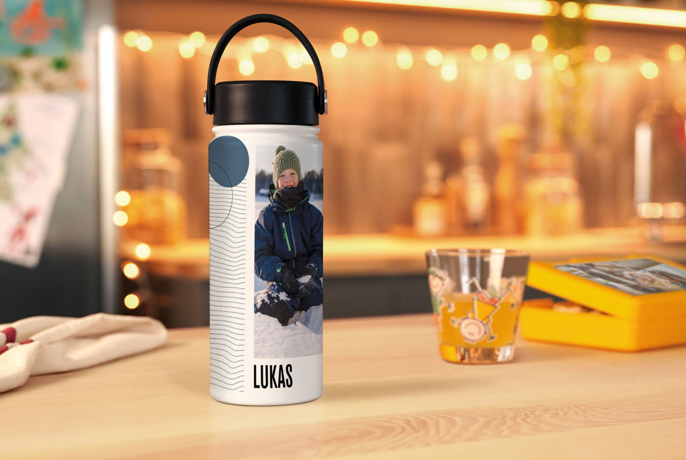 Personalisierte Edelstahl-Trinkflasche mit Foto eines Kindes im Winter und dem Namen „Lukas“ auf einem festlich beleuchteten Tisch.