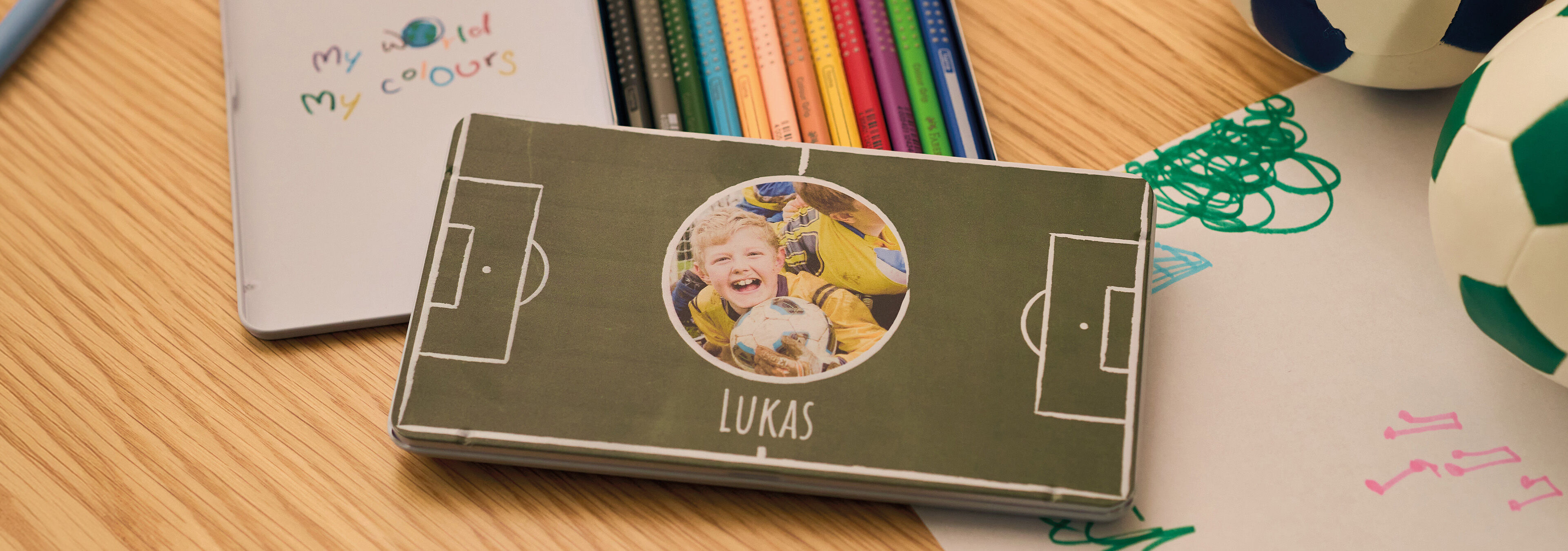 Personalisierte Stiftebox mit Kinderfoto und Fußballmotiv neben bunten Stiften auf einem Tisch.