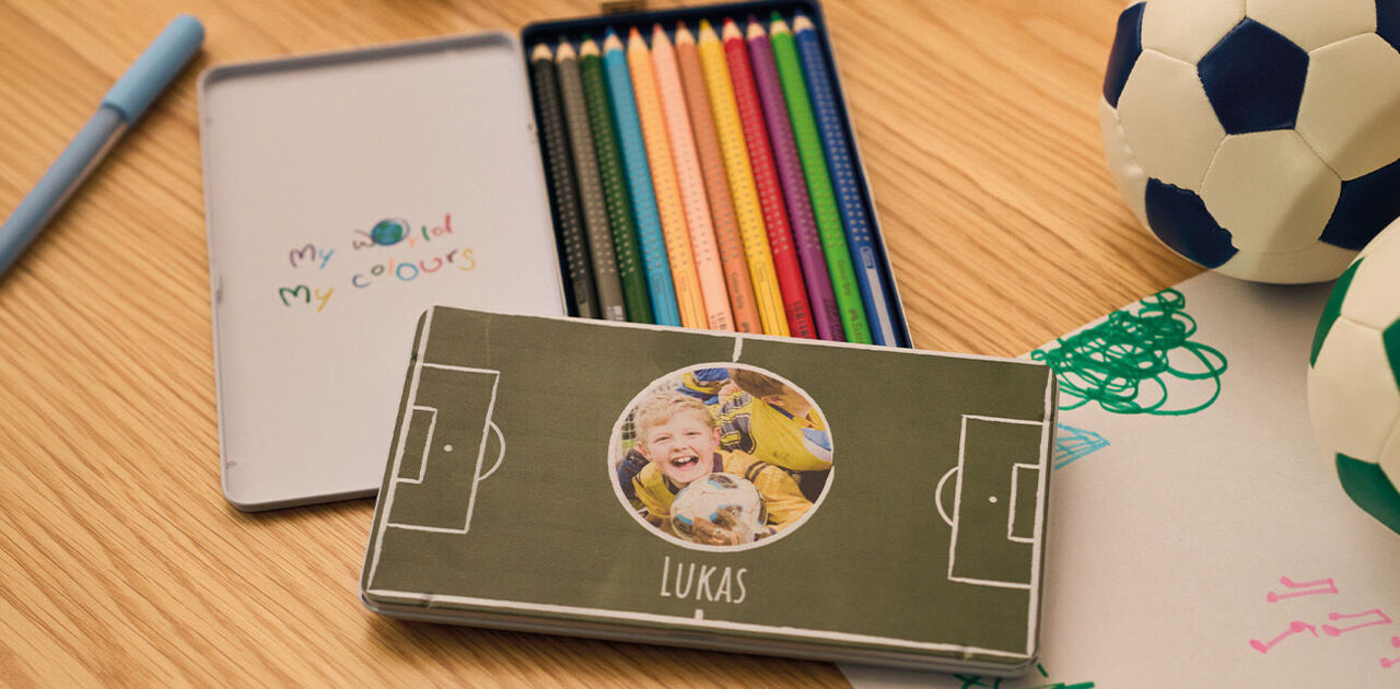 Personalisierte Stiftebox mit Kinderfoto und Fußballmotiv neben bunten Stiften auf einem Tisch.