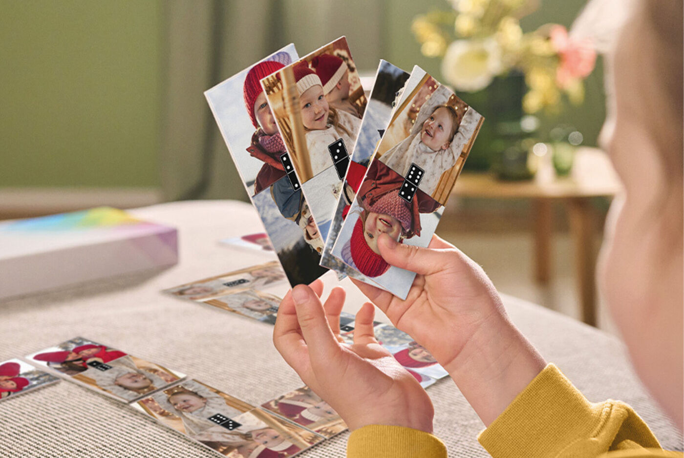 Kind hält mehrere Foto-Domino-Karten mit Kinderfotos in den Händen, im Hintergrund die bereits gelegten Karten.