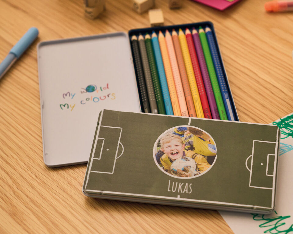 Personalisierte Stiftebox mit Kinderfoto und Fußballmotiv neben bunten Stiften auf einem Tisch.