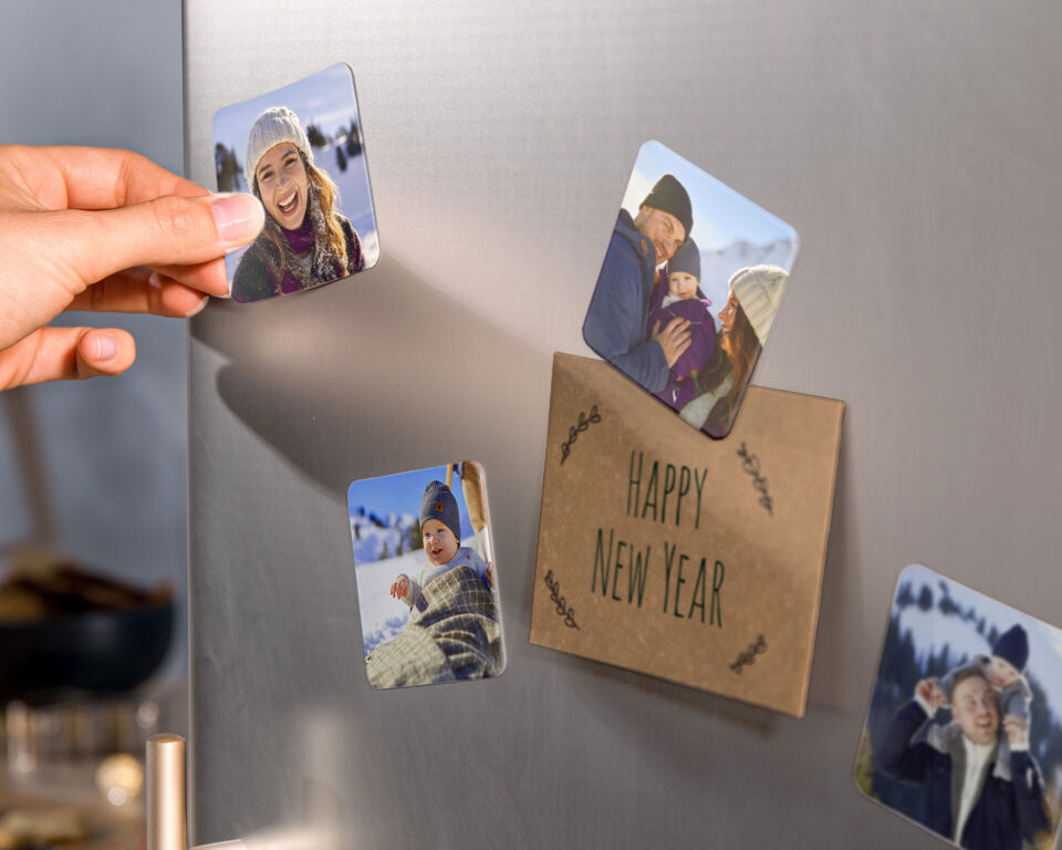 Person befestigt personalisierte Fotomagnete mit winterlichen Familienfotos an einem Kühlschrank neben einer Karte mit der Aufschrift „Happy