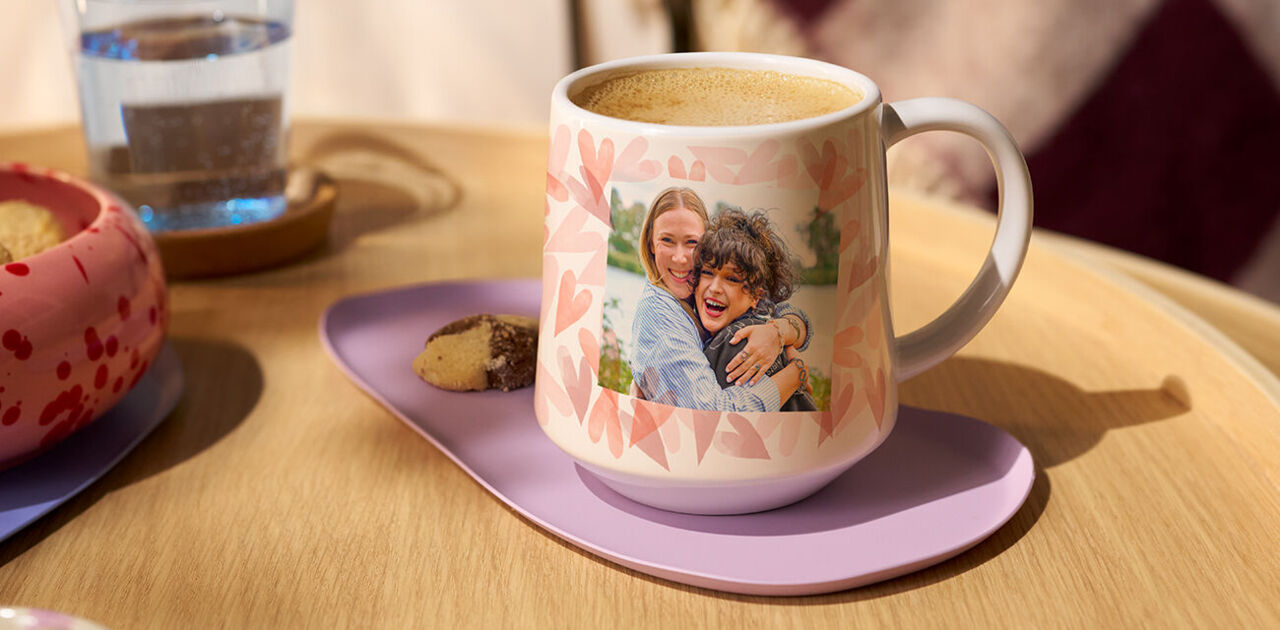 Personalisierte Tasse mit Foto von zwei lachenden Freundinnen auf einem Tisch mit Kaffee und Gebäck.